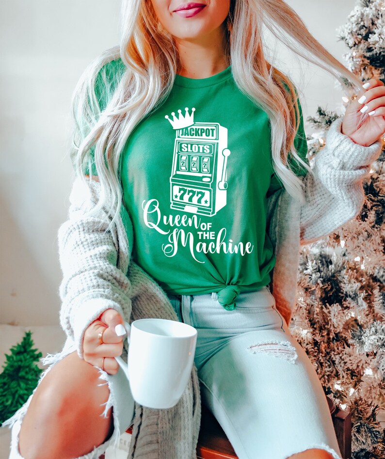 May include: A green t-shirt with a white graphic of a slot machine with the text "Jackpot Slots" and "Queen of the Machine".