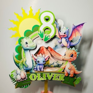Op de afbeelding: Een groene, gele en blauwe taarttopper met een zon, een vulkaan, het cijfer 8 en vier dinosaurussen. De dinosaurussen zijn een groene en blauwe dinosaurus, een paarse dinosaurus, een roze dinosaurus en een oranje dinosaurus. De taarttopper zegt "Oliver" op een groene banner.