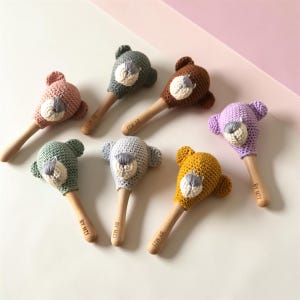 Puede incluir: Siete sonajeros de oso tejidos a ganchillo con mangos de madera, cada uno con una cabeza de diferente color: rosa, verde salvia, gris oscuro, marrón, gris claro, amarillo mostaza y lavanda. Cada mango tiene grabado el texto BY MZI.