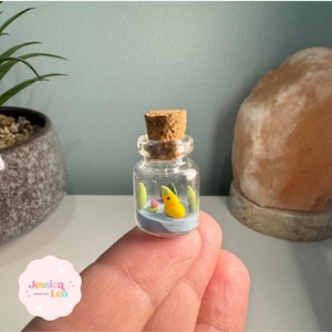 Peut inclure: Une petite bouteille en verre avec un bouchon en liège, contenant une scène miniature d'un oiseau jaune dans un étang bleu avec des plantes vertes.