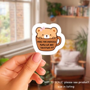 May include: A die-cut sticker showing a cartoon bear with a pink bow in a brown coffee mug. The mug has the text "DOES THE PROCESS KNOW WE ARE TRUSTING IT?" in brown. The sticker is held by a hand with pink nail polish.