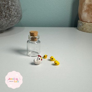 Puede incluir: Una botella de vidrio transparente con un tapón de corcho, junto a una figura de gallina blanca con cresta roja y tres figuras de pollitos amarillos. Las figuras están sobre una superficie blanca.