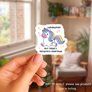 May include: A white sticker with a cartoon unicorn, rainbow mane and tail. The text on the sticker reads "UNDIAGNOSED BUT THERE'S DEFINITELY SOMETHING", with hearts and stars. The sticker is held in a person's hand.