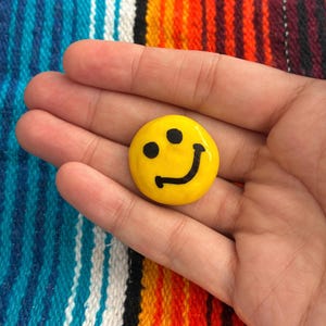 May include: A small, round, yellow stone featuring a hand-painted black smiley face with two circular eyes and a curved mouth. The surface of the stone is smooth and glossy, highlighting the vibrant yellow paint against the simple black facial features.