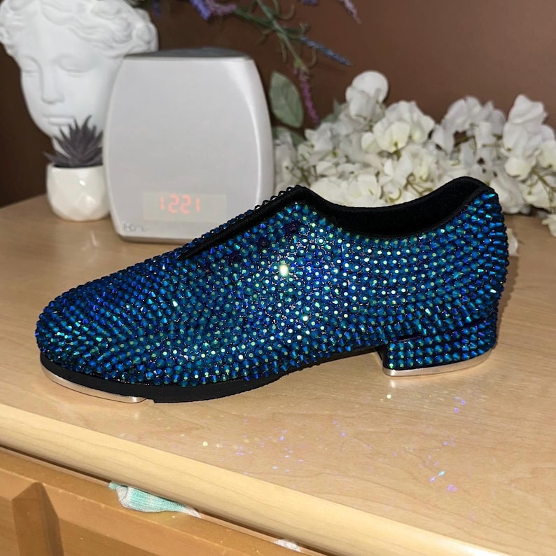 May include: A pair of blue dance shoes covered in iridescent rhinestones. The shoes have a black leather upper and a black sole with a small heel.