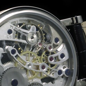 May include: Close-up of a silver-toned skeleton watch, showcasing intricate gears, jewels, and mechanical components. The watch face reveals the inner workings, with a black leather band. The text "Wandolec Art Studio" is visible.
