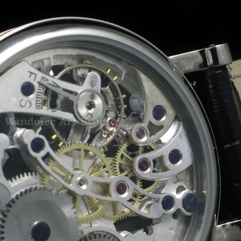 May include: Close-up of a silver-toned skeleton watch, showcasing intricate gears, jewels, and mechanical components. The watch face reveals the inner workings, with a black leather band. The text "Wandolec Art Studio" is visible.
