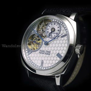 Puede incluir: Reloj de pulsera plateado con correa de cuero negro. La esfera del reloj presenta un patr&oacute;n plateado texturizado, agujas azules y n&uacute;meros romanos. El texto "DUAL TIME NEW YORK" es visible.