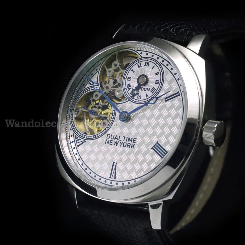 Puede incluir: Reloj de pulsera plateado con correa de cuero negro. La esfera del reloj presenta un patr&oacute;n plateado texturizado, agujas azules y n&uacute;meros romanos. El texto "DUAL TIME NEW YORK" es visible.