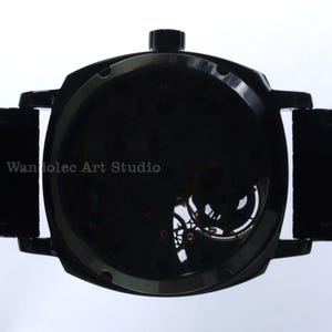 Puede incluir: Un reloj de pulsera negro con esfera esqueleto, que muestra los intrincados mecanismos internos. El reloj tiene una correa oscura y una corona en la parte superior. El texto "Wandolec Art Studio" es visible.