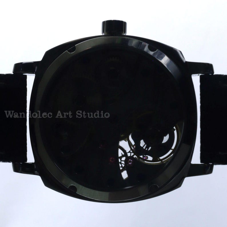 Puede incluir: Un reloj de pulsera negro con esfera esqueleto, que muestra los intrincados mecanismos internos. El reloj tiene una correa oscura y una corona en la parte superior. El texto "Wandolec Art Studio" es visible.
