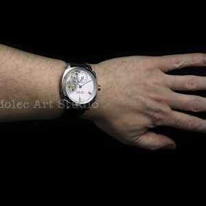 Puede incluir: Un reloj de pulsera plateado con esfera blanca y correa de cuero negro. El reloj presenta un complejo mecanismo interno visible a trav&eacute;s de una peque&ntilde;a ventana. El reloj est&aacute; en una mu&ntilde;eca sobre un fondo negro.