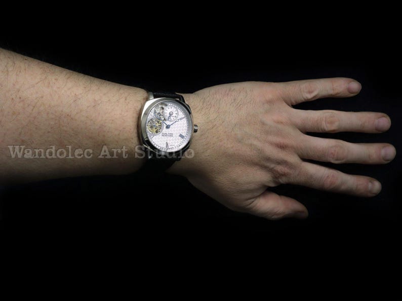 Puede incluir: Un reloj de pulsera plateado con esfera blanca y correa de cuero negro. El reloj presenta un complejo mecanismo interno visible a trav&eacute;s de una peque&ntilde;a ventana. El reloj est&aacute; en una mu&ntilde;eca sobre un fondo negro.