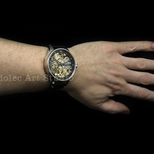 May include: A silver-toned wristwatch with a black leather band. The watch face is transparent, revealing the intricate gold-colored mechanical movement. The watch is on a wrist against a black background.