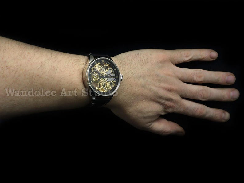May include: A silver-toned wristwatch with a black leather band. The watch face is transparent, revealing the intricate gold-colored mechanical movement. The watch is on a wrist against a black background.