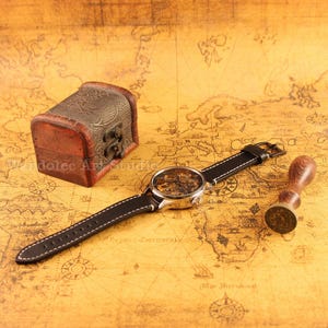 May include: A vintage-style watch with a skeleton dial and black leather strap is displayed next to a small wooden chest and a wooden stamp. The watch has a silver-colored case and a detailed inner mechanism.