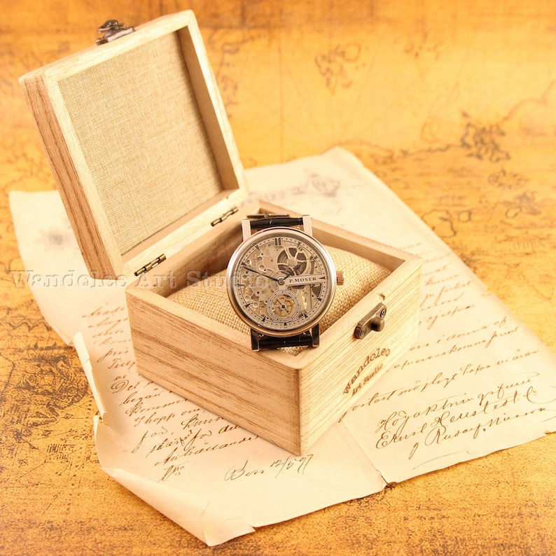 May include: A vintage-style wristwatch with a skeleton dial, displayed in an open wooden box lined with beige fabric. The watch has a black leather band and is set against a backdrop of aged paper.