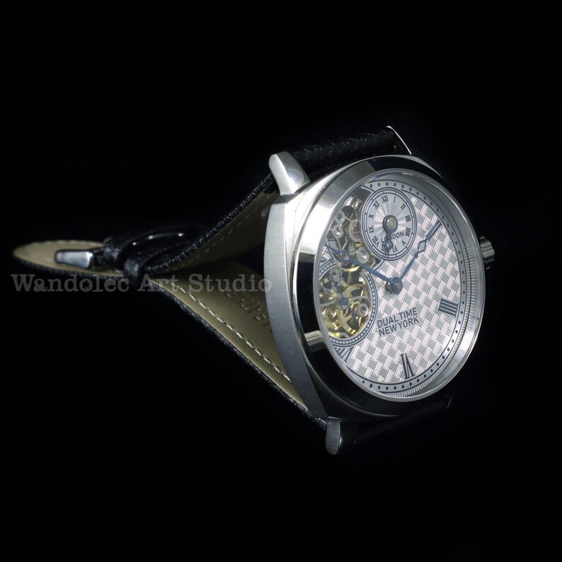 Puede incluir: Un reloj de pulsera plateado de doble huso horario con una correa de cuero negro. La esfera del reloj tiene un patr&oacute;n de cuadros con n&uacute;meros romanos y dos esferas m&aacute;s peque&ntilde;as. El texto "DUAL TIME NEW YORK" es visible.