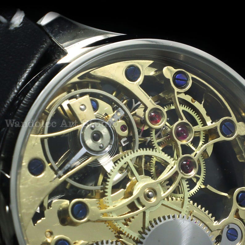 May include: Close-up of a skeleton watch with a black leather band. The watch face reveals intricate gold-colored gears, ruby jewels, and blue accents. The watch has a silver-colored bezel and a partially visible black band.