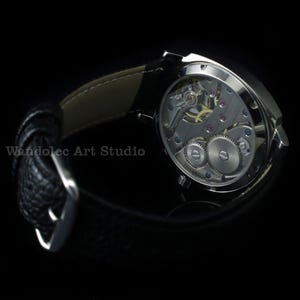 Puede incluir: Primer plano de un reloj de pulsera plateado con una correa de cuero negro. La parte trasera del reloj es visible, mostrando los intrincados mecanismos internos. El texto "Wandolec Art Studio" es visible. Fondo negro.