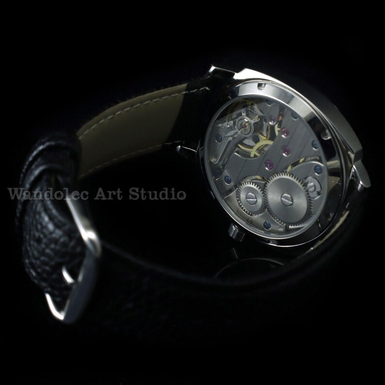 Puede incluir: Primer plano de un reloj de pulsera plateado con una correa de cuero negro. La parte trasera del reloj es visible, mostrando los intrincados mecanismos internos. El texto "Wandolec Art Studio" es visible. Fondo negro.