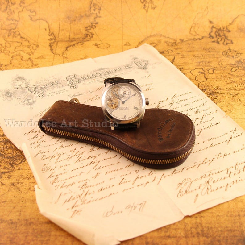 Puede incluir: Un reloj plateado con una correa de cuero negro descansa sobre un estuche de cuero marr&oacute;n. El reloj tiene una esfera blanca con engranajes visibles. El estuche tiene cremallera y est&aacute; sobre papel envejecido con escritura manuscrita.