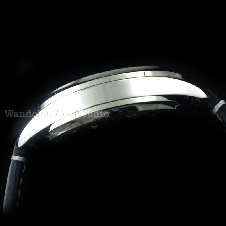 May include: Close-up of a silver wristwatch with a black leather band. The watch face is not visible. The band is stitched with white thread. The watch is set against a black background. The text "Wandelco Art Studio" is visible.