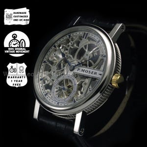 Op de afbeelding: Een zilverkleurig skeleton horloge met een zwarte leren band. De wijzerplaat van het horloge heeft ingewikkelde tandwielen en de tekst "P.MOSER". Het heeft een goudkleurige kroon en de tekst "HANDMADE CUSTOMIZED ONE-OF-KIND" is zichtbaar.