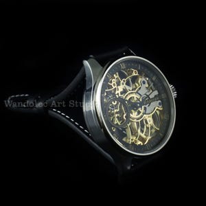 May include: A silver-toned skeleton watch with a black face and a black leather band. The watch face features visible gears and Roman numeral hour markers. The band is stitched with white thread. The text "Wandolec Art Studio" is visible.