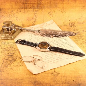 May include: A vintage-style watch with a black leather band and a silver case, showcasing the intricate inner workings. The watch rests on a parchment-like paper, alongside a feather quill, inkwell, and eyeglasses, suggesting a theme of history and craftsmanship.
