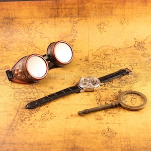 May include: Steampunk-style goggles with copper-colored frames and mirrored lenses, a skeleton watch with a black leather band, and a brass magnifying glass. The items are arranged on a vintage map.