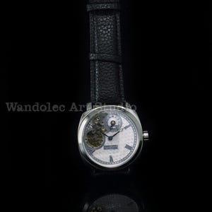 Puede incluir: Un reloj de pulsera plateado con una correa de cuero negro. La esfera del reloj es blanca con intrincados detalles mec&aacute;nicos y el texto "REAL TIME NEW YORK". El reloj est&aacute; sobre un fondo negro.