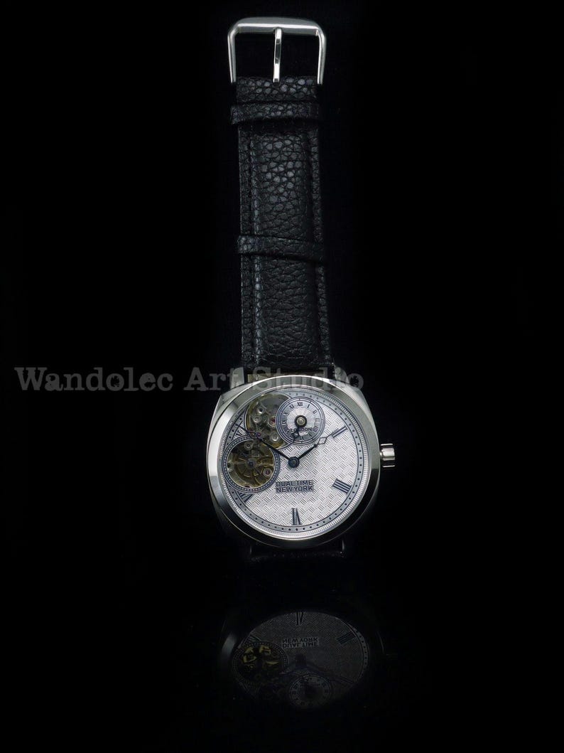 Puede incluir: Un reloj de pulsera plateado con una correa de cuero negro. La esfera del reloj es blanca con intrincados detalles mec&aacute;nicos y el texto "REAL TIME NEW YORK". El reloj est&aacute; sobre un fondo negro.