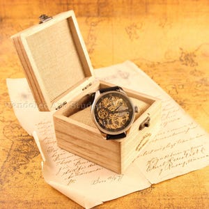 May include: A mechanical wristwatch with a black leather band rests in an open wooden box lined with beige fabric. The watch has a silver case and a visible intricate movement. The box sits on a vintage map and aged paper.