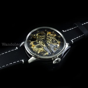 May include: A silver-toned skeleton watch with a black face and gold-colored internal mechanisms. The watch has Roman numeral hour markers and a black leather band with white stitching. The watch is a luxury item.