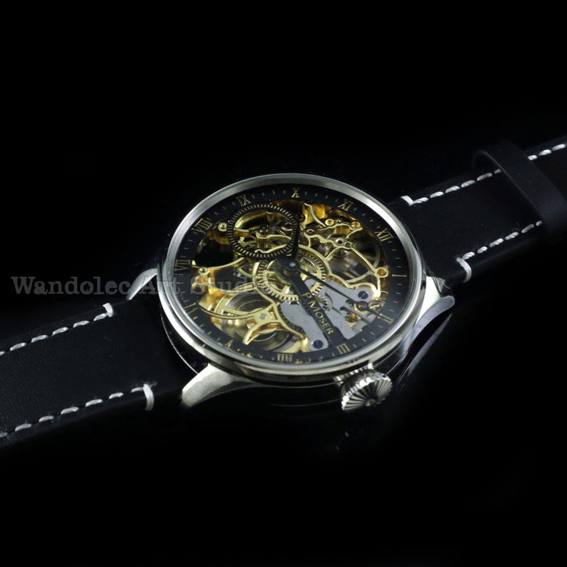 May include: A silver-toned skeleton watch with a black face and gold-colored internal mechanisms. The watch has Roman numeral hour markers and a black leather band with white stitching. The watch is a luxury item.