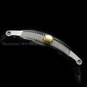 May include: A silver-toned watch band with a ribbed center section and a gold-colored circular accent. The band has silver end pieces with small circular details. The text "Wandolec Art Studio" is visible.