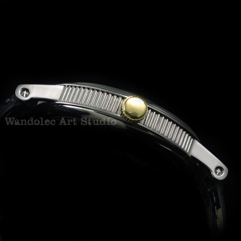 May include: A silver-toned watch band with a ribbed center section and a gold-colored circular accent. The band has silver end pieces with small circular details. The text "Wandolec Art Studio" is visible.