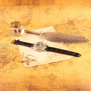 May include: A vintage-themed still life featuring a skeleton watch with a black leather band, an inkwell, a quill pen, and round eyeglasses. The items rest on a map-printed surface, creating an antique aesthetic. The watch face is visible, showcasing its intricate inner workings.