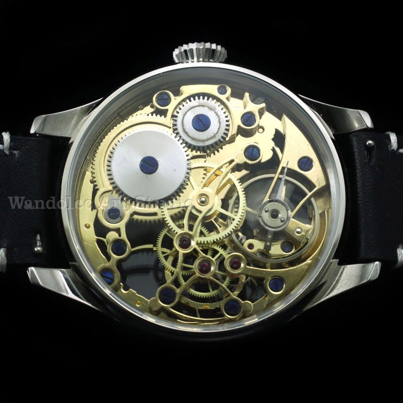 May include: A silver-toned wristwatch with a transparent face revealing intricate gold-colored gears, blue accents, and red jewels. The watch has a black leather band with white stitching. The watch face is approximately 1.75 inches in diameter.