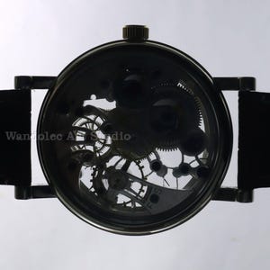 May include: A black skeleton watch with visible gears and a leather strap. The watch face displays intricate mechanical components. The text "Wanolec Art Studio" is visible on the watch face.