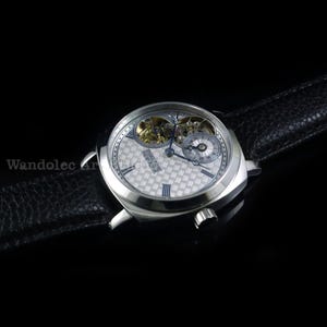 Puede incluir: Un reloj de pulsera plateado con una esfera estampada y una correa de cuero negro. El reloj presenta dos esferas m&aacute;s peque&ntilde;as y mecanismos internos visibles. El reloj se encuentra sobre un fondo negro, resaltando sus detalles.