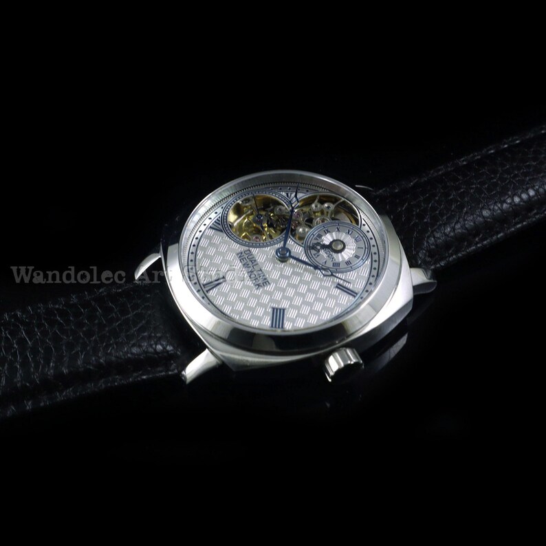 Puede incluir: Un reloj de pulsera plateado con una esfera estampada y una correa de cuero negro. El reloj presenta dos esferas m&aacute;s peque&ntilde;as y mecanismos internos visibles. El reloj se encuentra sobre un fondo negro, resaltando sus detalles.