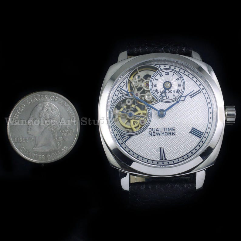 Puede incluir: Un reloj de pulsera plateado con esfera blanca y correa de cuero negro. El reloj presenta una pantalla de doble hora y n&uacute;meros romanos. Una moneda est&aacute; al lado del reloj para la escala. El texto "DUAL TIME NEW YORK" es visible.