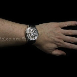 May include: A silver skeleton watch with a black leather band is displayed on a wrist. The watch face reveals the intricate inner workings of the timepiece. The background is solid black.