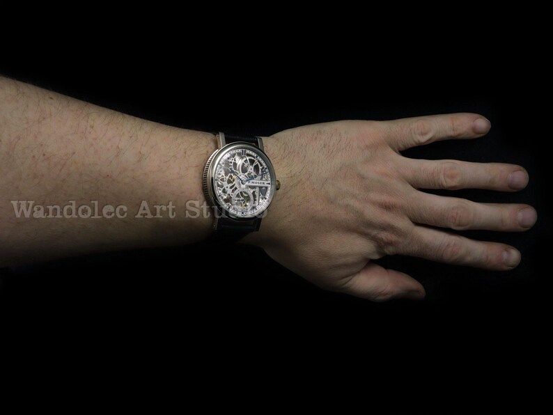 May include: A silver skeleton watch with a black leather band is displayed on a wrist. The watch face reveals the intricate inner workings of the timepiece. The background is solid black.
