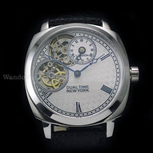 Puede incluir: Reloj de pulsera plateado con esfera texturizada y agujas azules. El reloj presenta dos esferas m&aacute;s peque&ntilde;as y el texto "DUAL TIME NEW YORK". Tiene una correa de cuero negro.