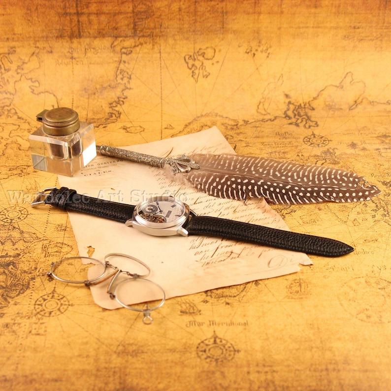 Puede incluir: Naturaleza muerta de inspiraci&oacute;n vintage con un reloj esqueleto plateado con correa de cuero negro, un tintero, una pluma y gafas redondas. Los objetos descansan sobre un papel con forma de mapa, creando una est&eacute;tica antigua.