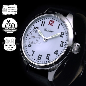 May include: A silver-toned Cortébert watch with a white face, black numerals, and a small seconds dial. The watch has a black leather band and a textured crown. The text "Cortébert" is printed on the watch face. The watch is labeled as handmade and vintage.