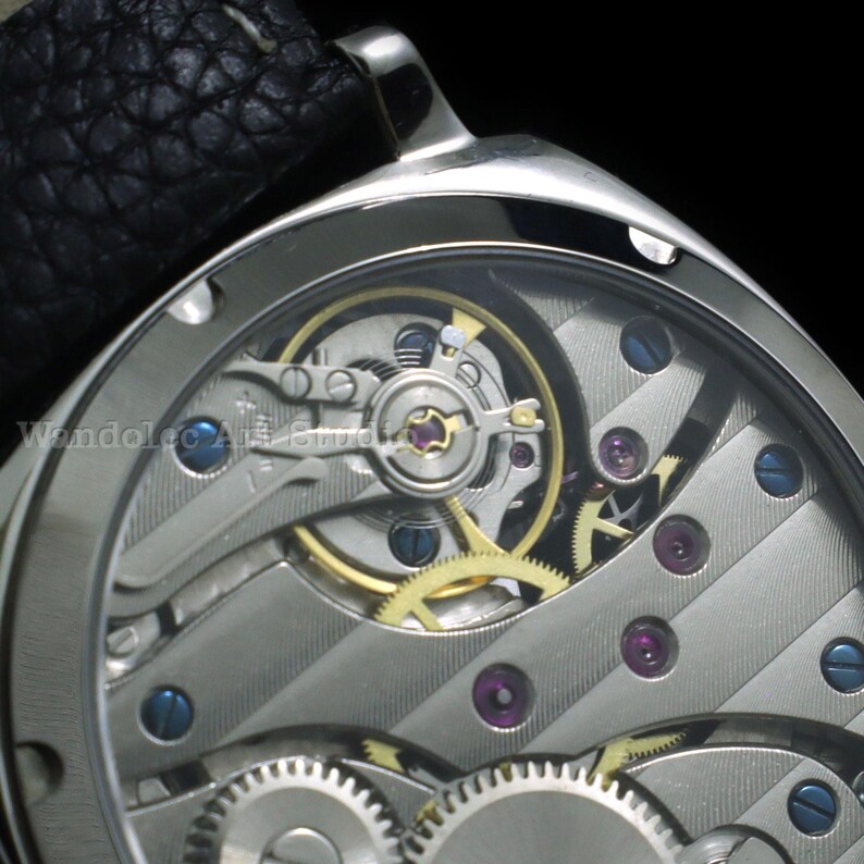 Puede incluir: Primer plano de un reloj de pulsera plateado con una correa de cuero negro. La esfera del reloj es transparente, revelando los intrincados mecanismos internos, incluyendo engranajes, joyas y muelles. El reloj tiene una caja de metal pulido.